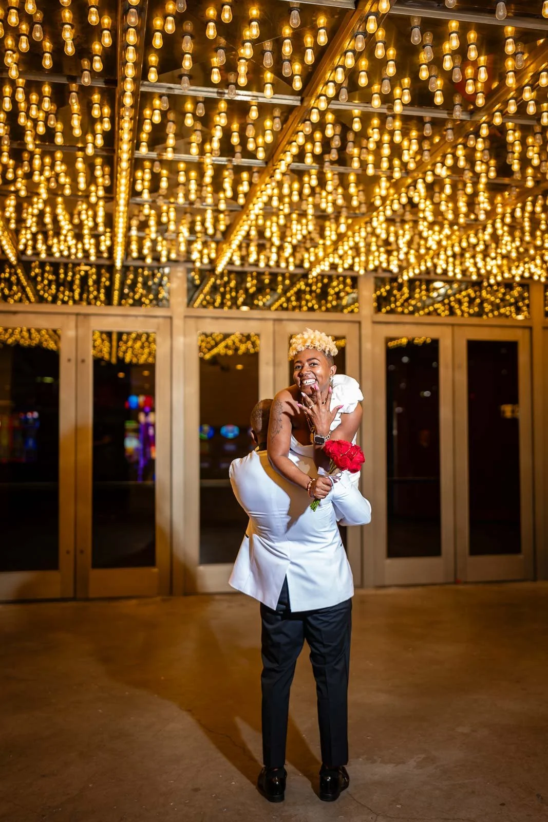 A joyful couple in wedding attire sharing a dance under a ceiling of warm light bulbs, with a woman holding a bouquet of red flowers.