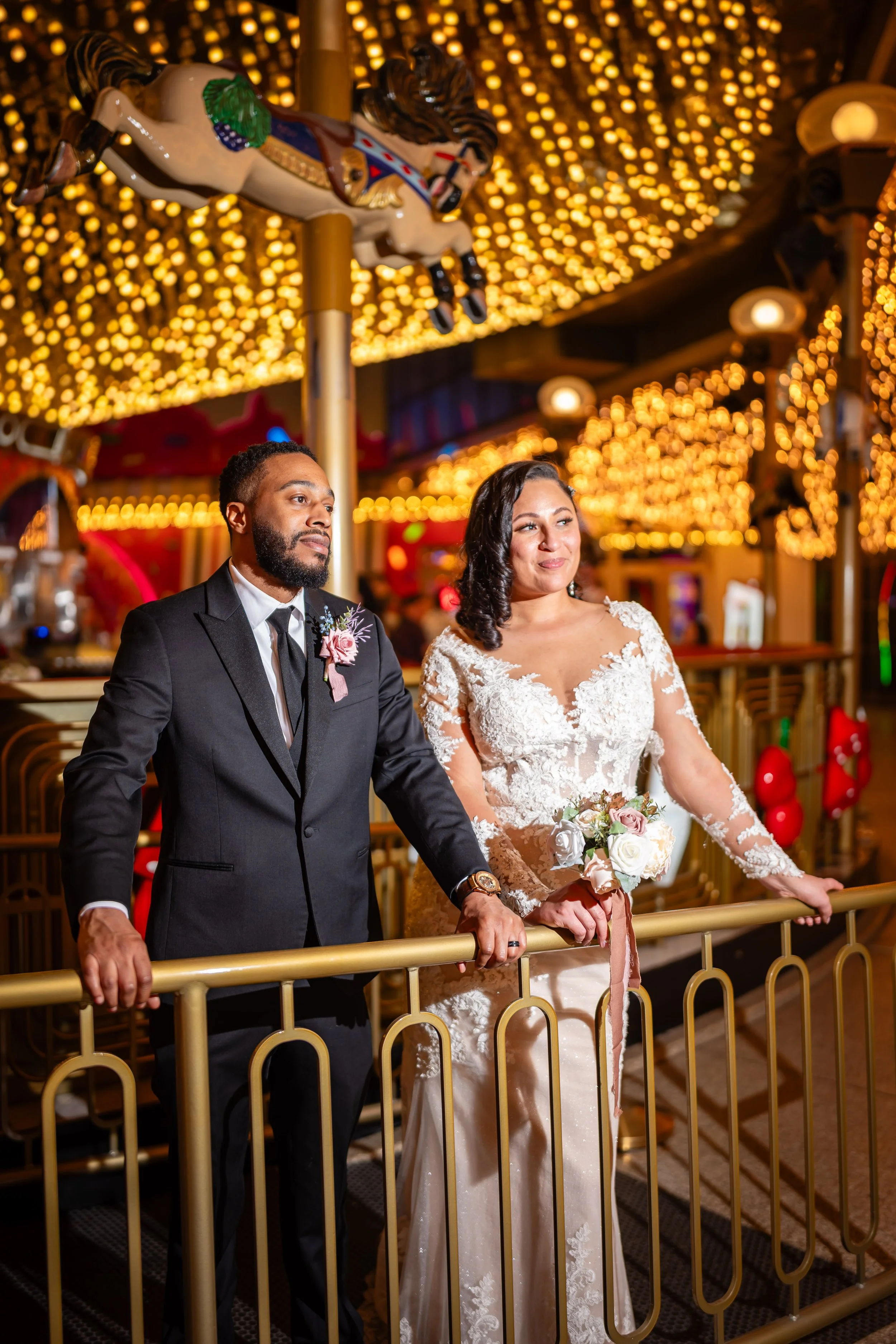 A bride and groom holding hands on a carousel ride at an amusement park, decorated with lights and horse figures.