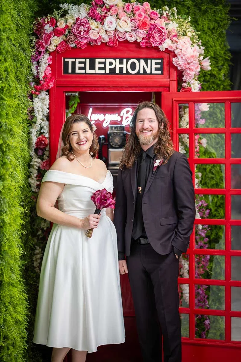 A bride and groom are smiling in front of a red telephone booth decorated with pink and white flowers, with greenery on the sides.