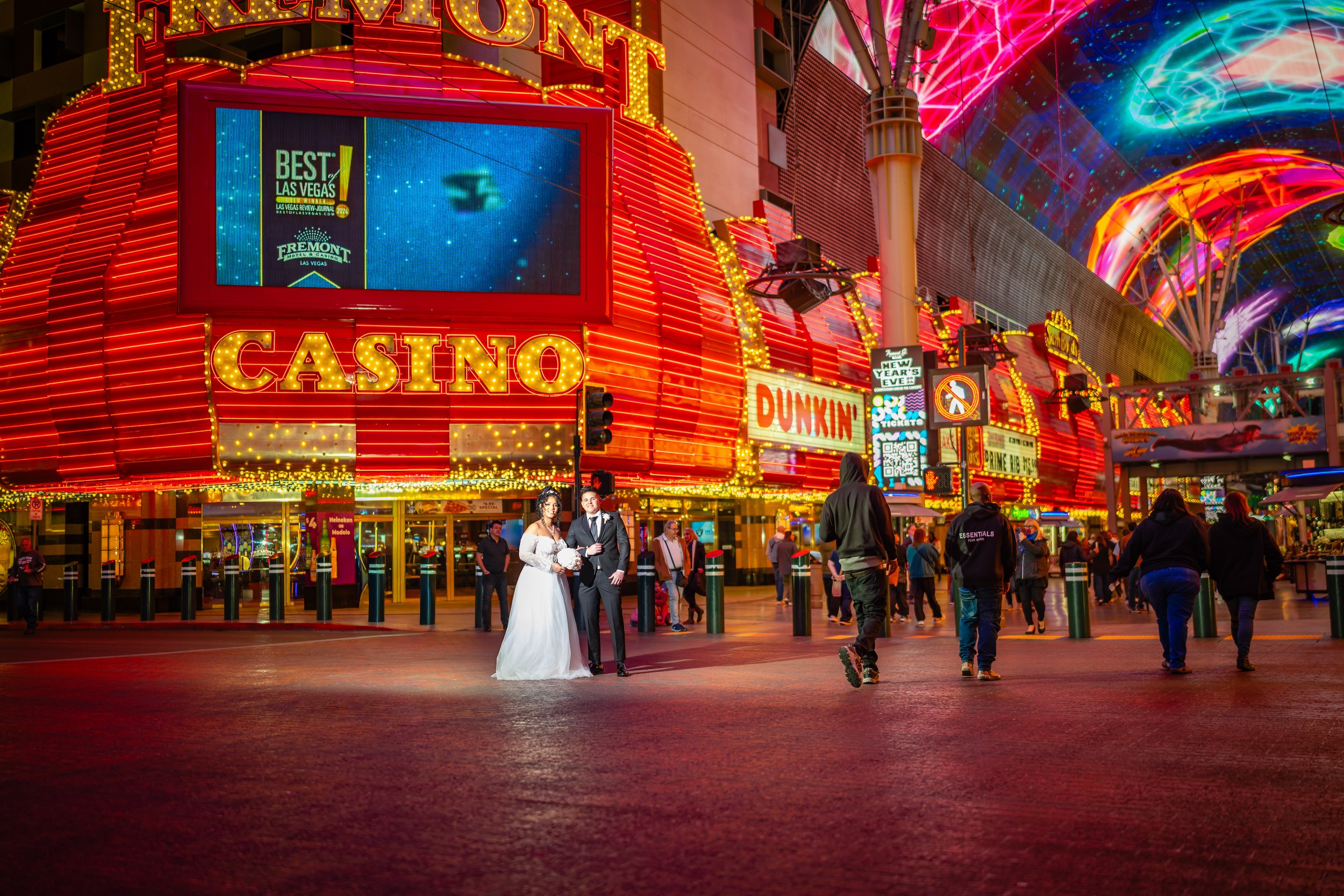 Brightly lit Las Vegas Strip at night featuring a casino entrance with a large red neon sign, a couple in wedding attire standing in front, and several pedestrians walking by, with colorful neon lights and a decorated ceiling overhead.