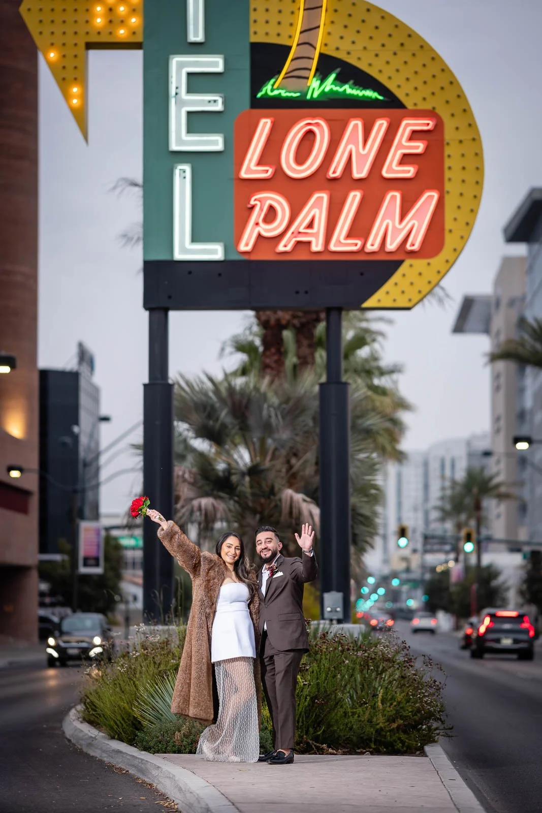 A couple dressed in wedding attire standing on a city sidewalk with a large neon sign reading 'LONE PALM' and 'HOTEL' behind them. The woman is holding a bouquet and wearing a white dress with a fur coat, while the man is in a suit. They are smiling 