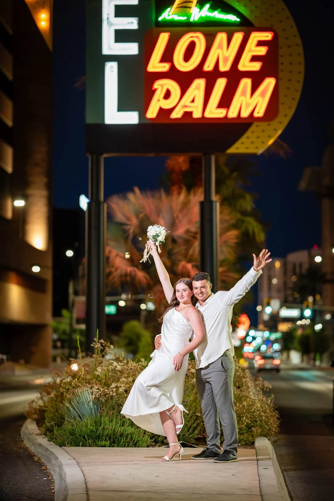A couple in wedding attire celebrating on a city street at night under a neon sign that reads "LONE PALM." The woman is holding a bouquet and raising her arm, while the man has his arm around her and is smiling. The background features palm trees and