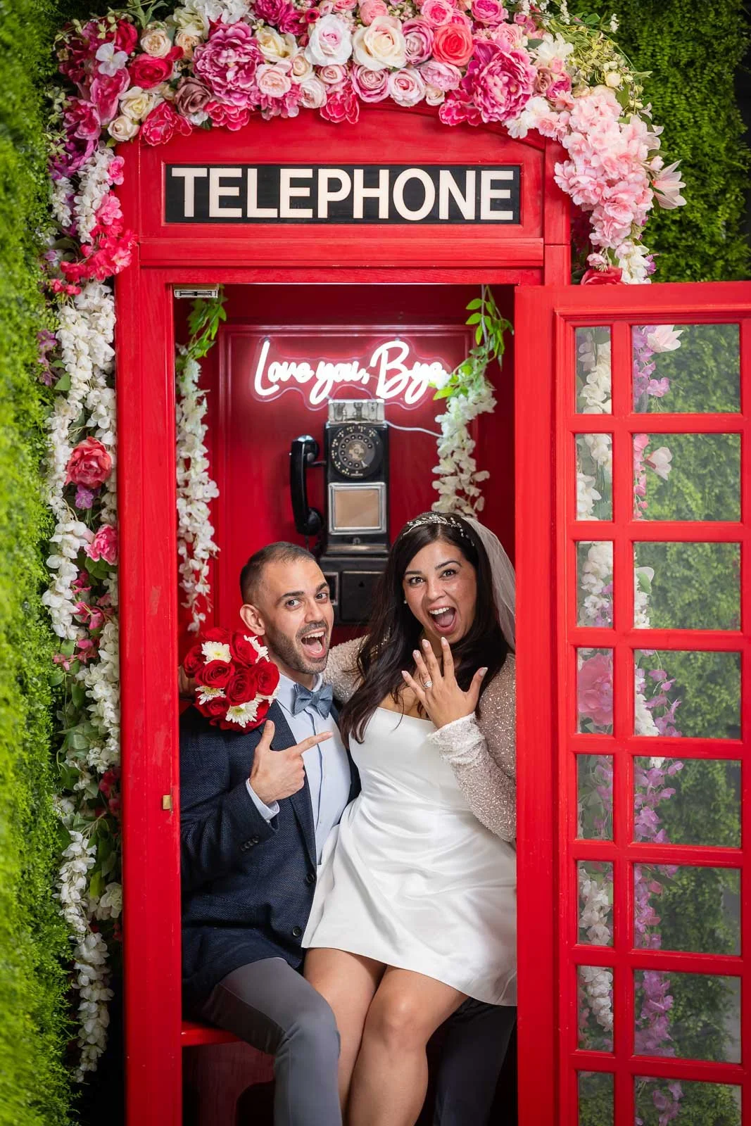 A bride and groom are inside a red vintage telephone booth decorated with pink and white flowers. The groom, pointing and smiling, holds a bouquet of red and white flowers. The bride, showing her wedding ring, has an excited expression. Behind them, 