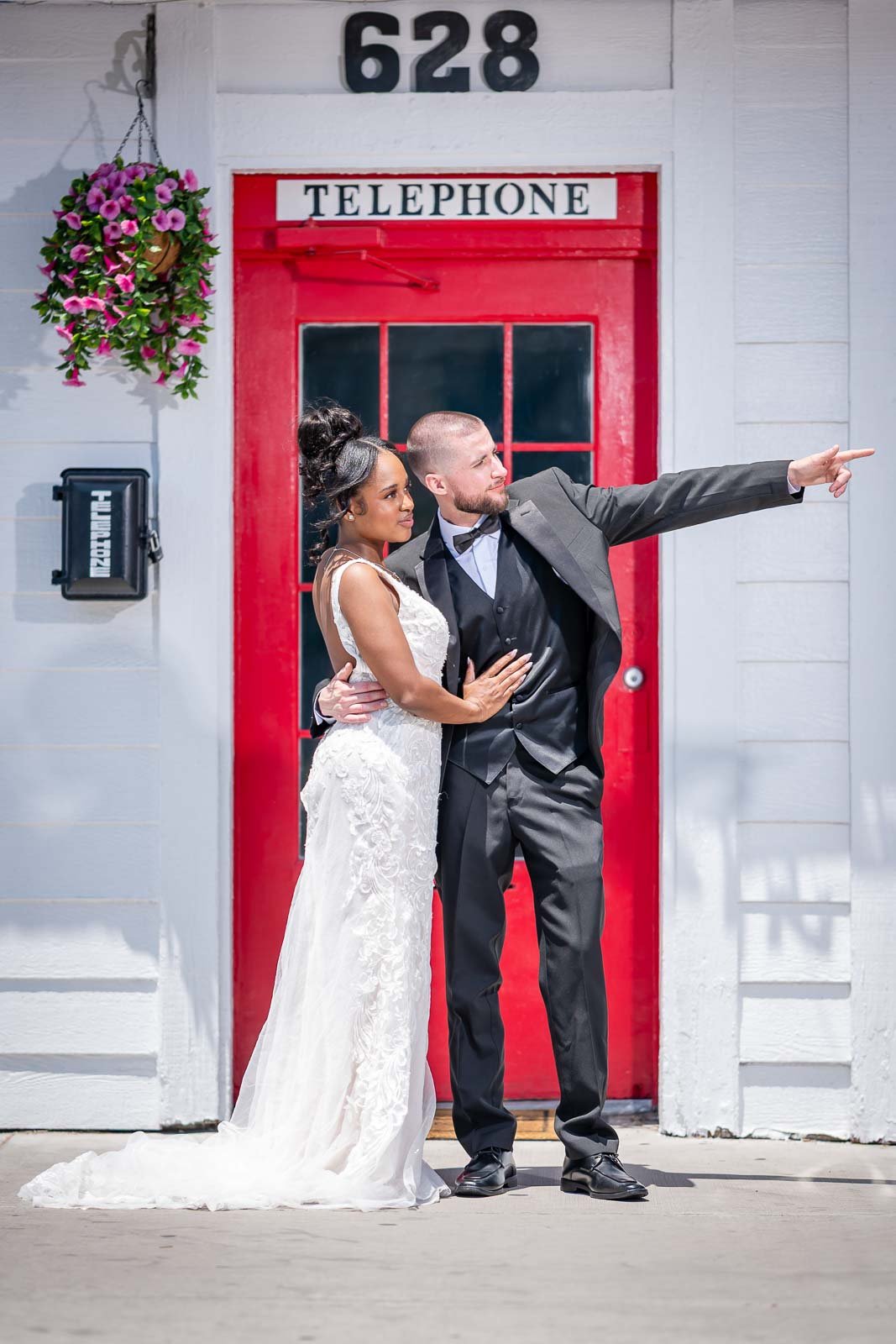 A newlywed couple standing in front of a red door with the word PETITION written above it. The man is pointing to the right while the woman leans into him, both dressed in wedding attire. The scene includes the house number 628 above the door and a hanging flower basket to the left.