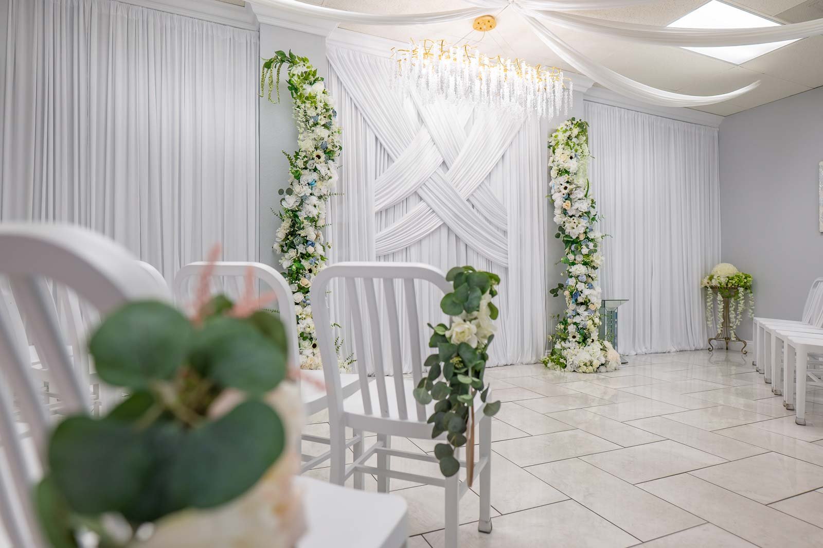 A decorated wedding ceremony space with white drapery, floral arrangements, and white chairs, ready for a wedding ceremony.
