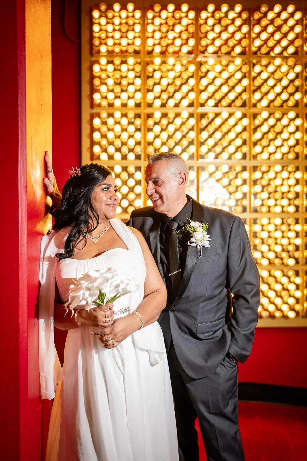 A bride and groom sharing a moment at their wedding, with the bride holding calla lilies and wearing a white dress, and the groom in a black tuxedo, standing against a background of warm glowing lights.