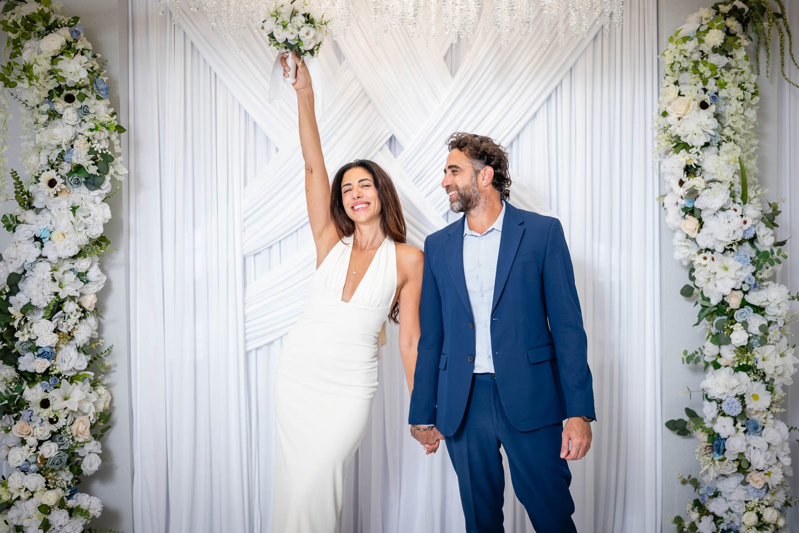 A woman in a white wedding dress holding a bouquet up in celebration, standing beside a man in a blue suit, both smiling, in front of a white floral and fabric wedding backdrop.