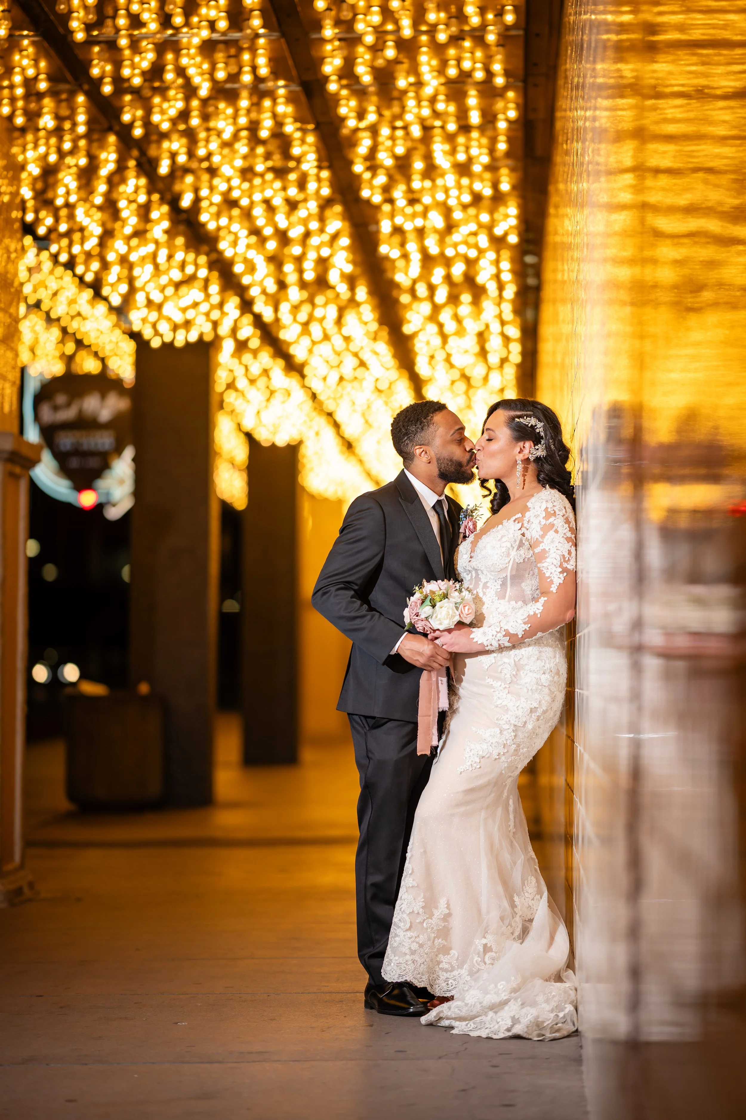 A newlywed couple sharing a kiss at night, with warm golden lights overhead and a soft reflection on a wall.