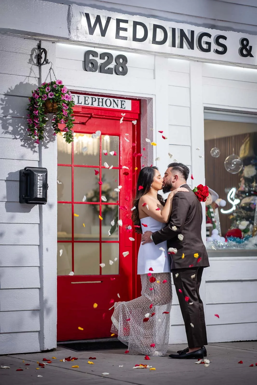 A newlywed couple sharing a kiss outside the Say I Do Chapel with a red telephone door, surrounded by falling rose and flower petals.