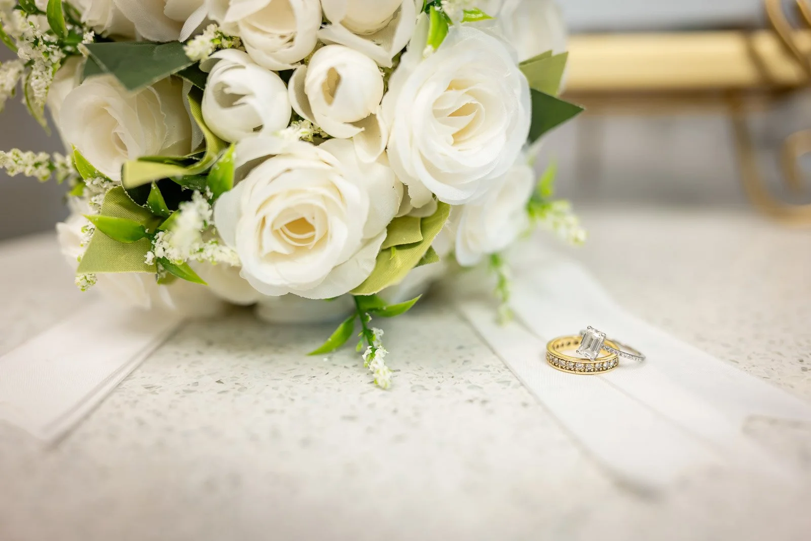A bridal bouquet of white roses and greenery, with two wedding rings, one gold and one silver, resting on a white ribbon on a textured surface.