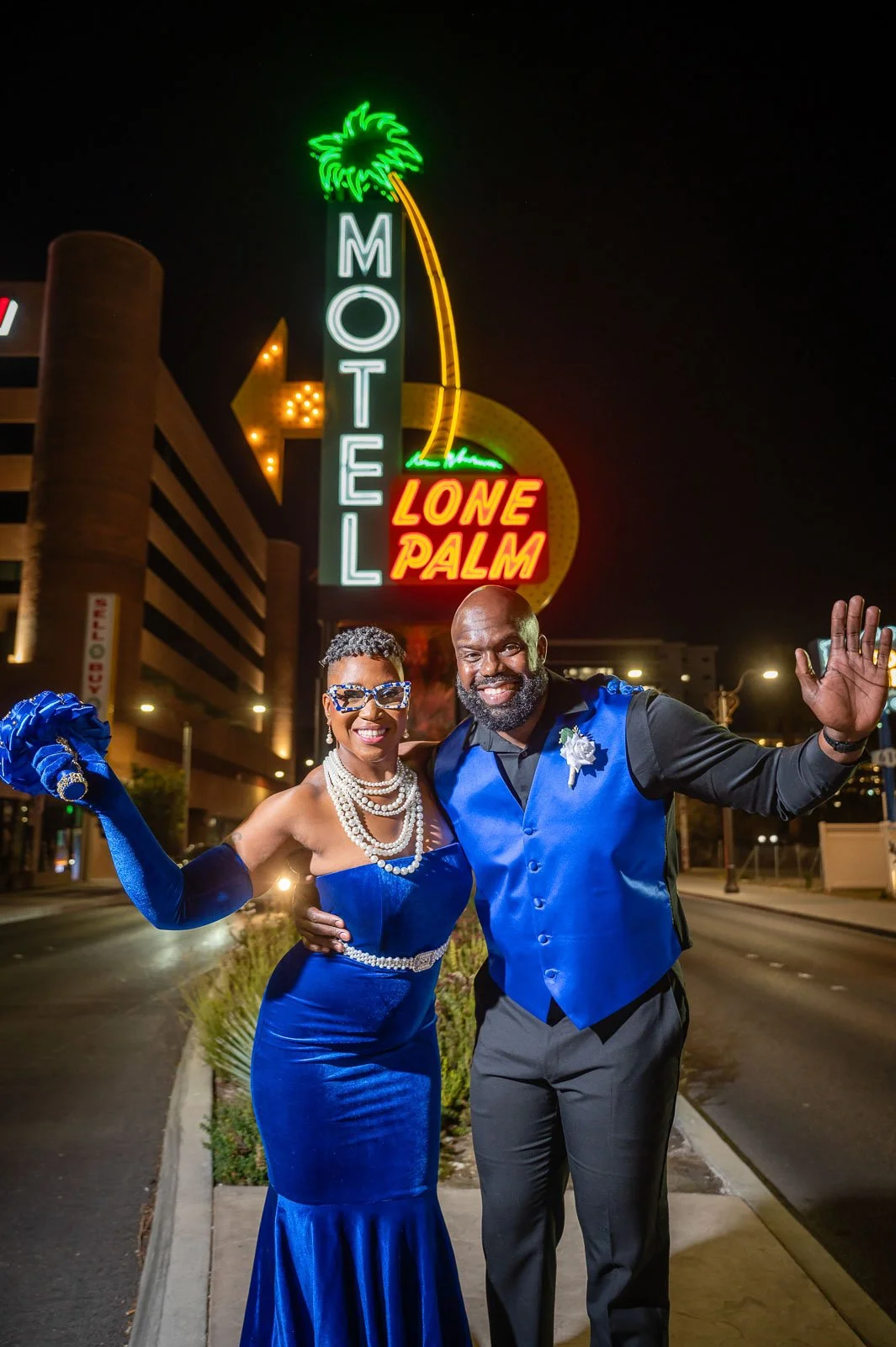 Photograph of a smiling couple dressed in formal blue attire posing in front of the Neon Motel sign at night.