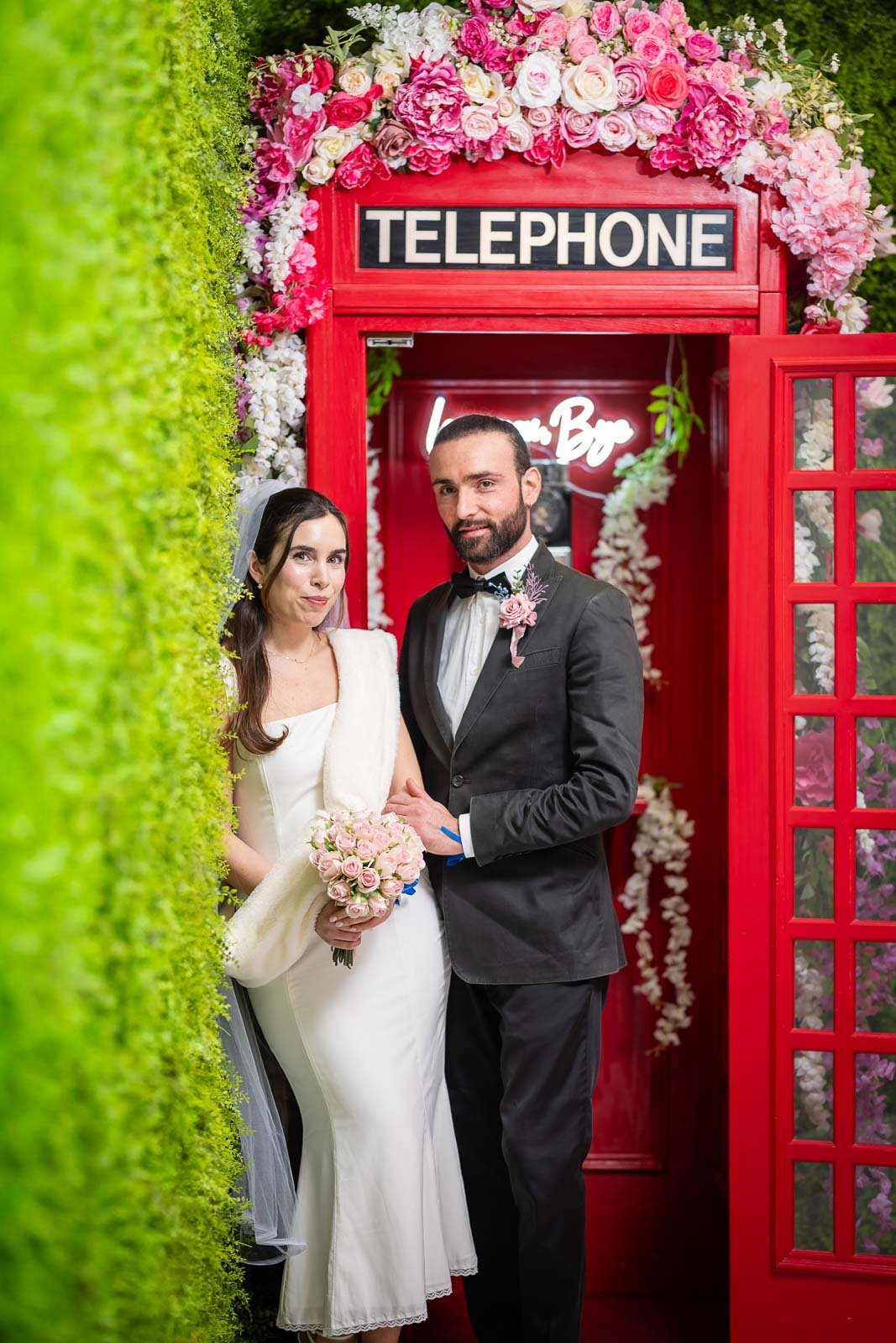 A newlywed couple standing inside a vintage red telephone booth decorated with a large arrangement of pink and white flowers. The bride is wearing a white dress and holding a bouquet of pink roses, while the groom is in a black tuxedo with a pink bou