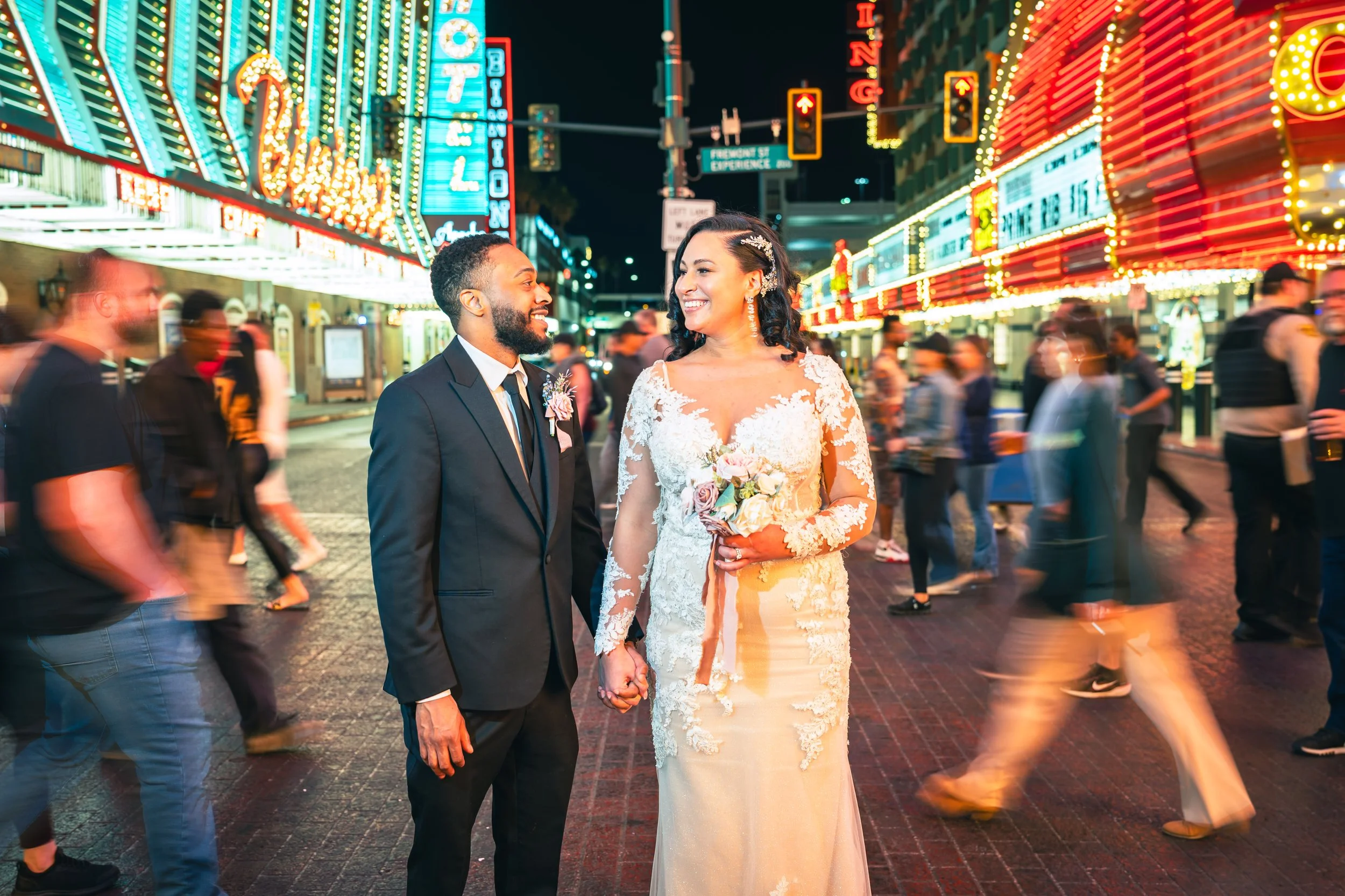 A bride and groom holding hands in a busy neon-lit street at night, smiling at each other.