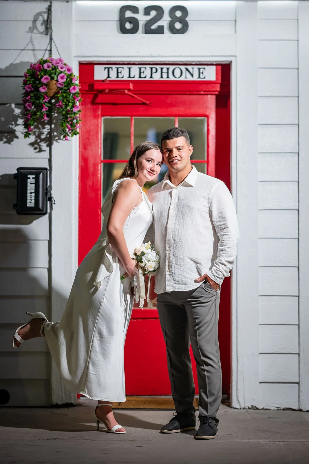 A couple standing in front of a red door with a white sign that says 'TELEPHONE'. The woman is wearing a white dress and holding a bouquet of white flowers, and the man is dressed in a white shirt and gray pants. The woman has one foot lifted behind 