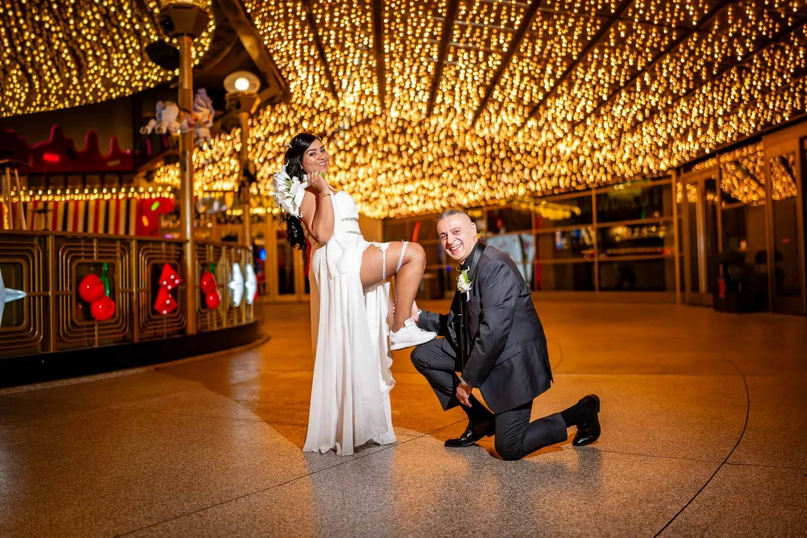 A bride and groom pose playfully under a ceiling covered with string lights in an amusement park or carnival setting, with festive decorations on a nearby railing in downtown Las Vegas.