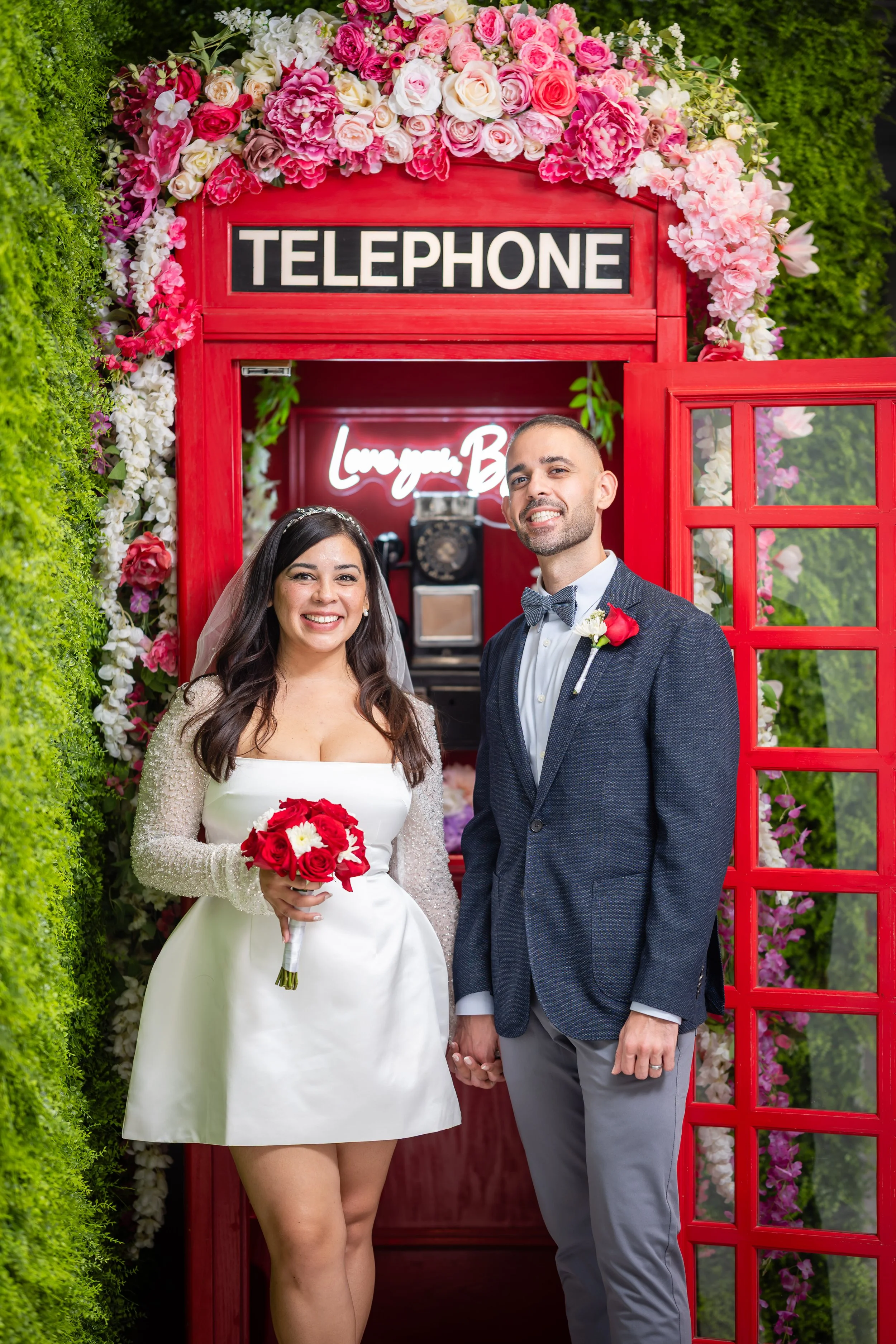 A bride and groom pose in front of a red telephone booth decorated with pink and white flowers at their wedding. The bride wears a white dress with lace sleeves and holds a bouquet of red roses with white flowers. The groom wears a dark blue blazer, 