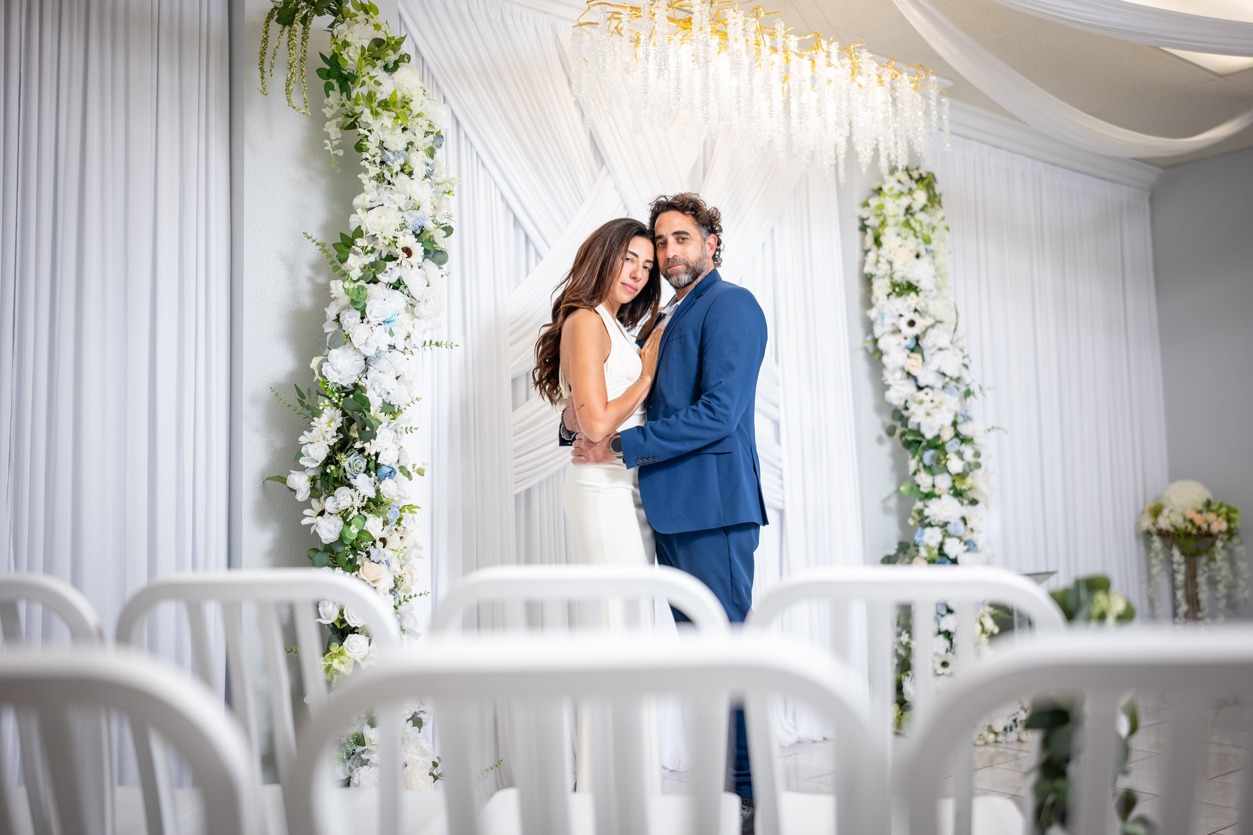 A couple dressed in wedding attire standing close together during their wedding ceremony, surrounded by white floral arrangements and drapery.