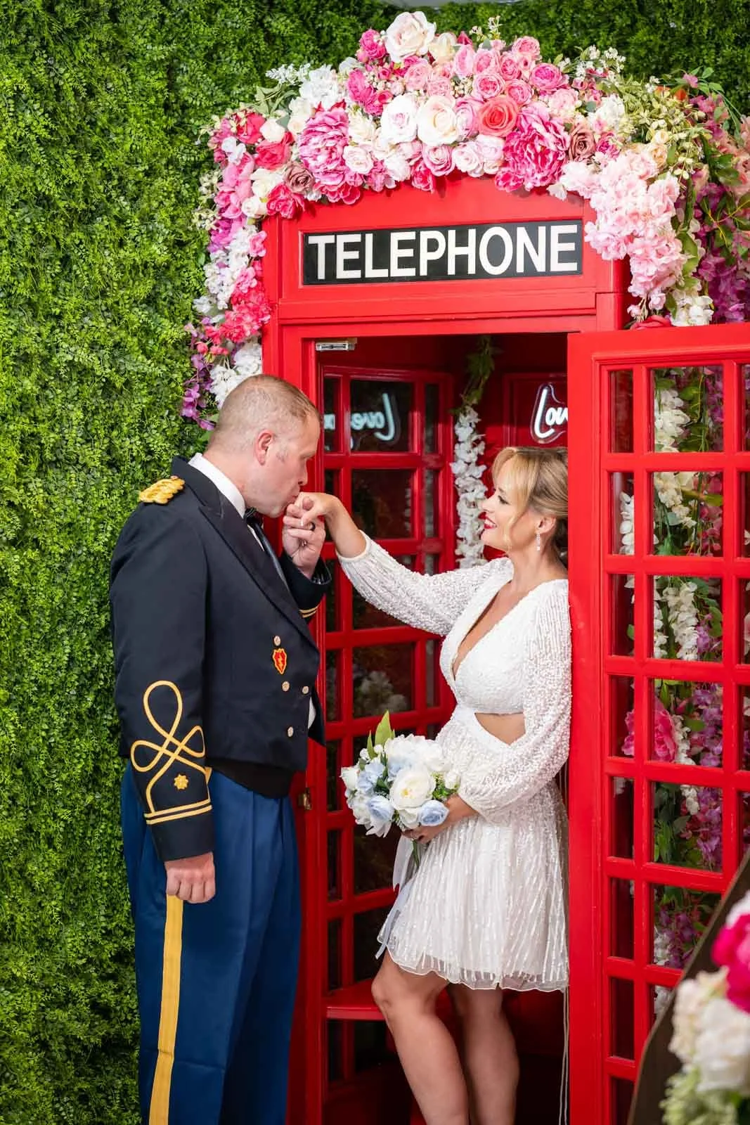 A woman in a white dress is touching a kiss on the lips of a man in a military uniform inside a red telephone booth decorated with pink and white flowers, with greenery on the left side.