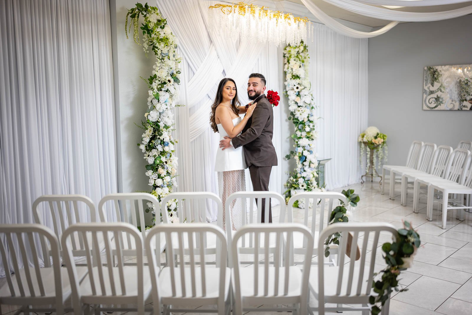A bride and groom stand together inside the Say I Do Chapel, surrounded by white chairs, with an arch decorated with white and green flowers and draped white fabric behind them.