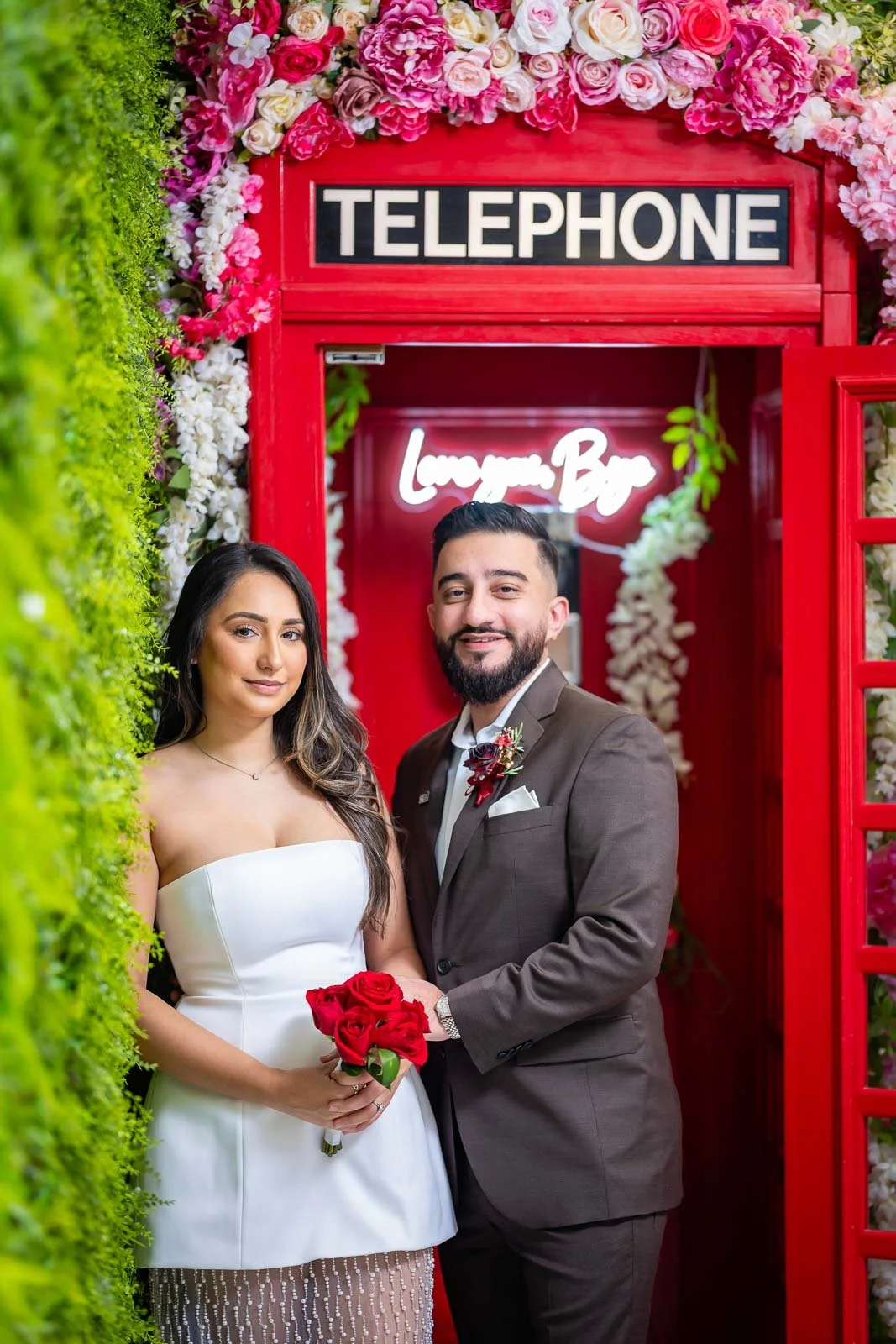 A bride and groom standing in front of a red telephone booth inside the Say I Do Chapel decorated with pink and white flowers. The bride holds a bouquet of red roses, and the groom wears a suit with a boutonniere.