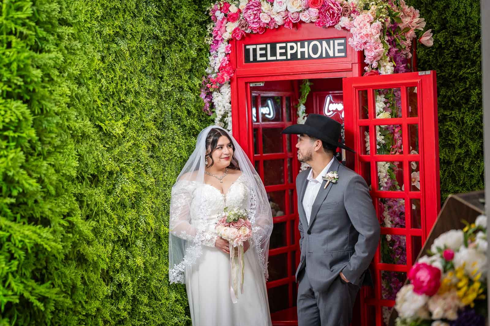 Bride and groom standing in front of a red telephone booth inside the Say I Do Chapel decorated with flowers, looking at each other during a wedding celebration.