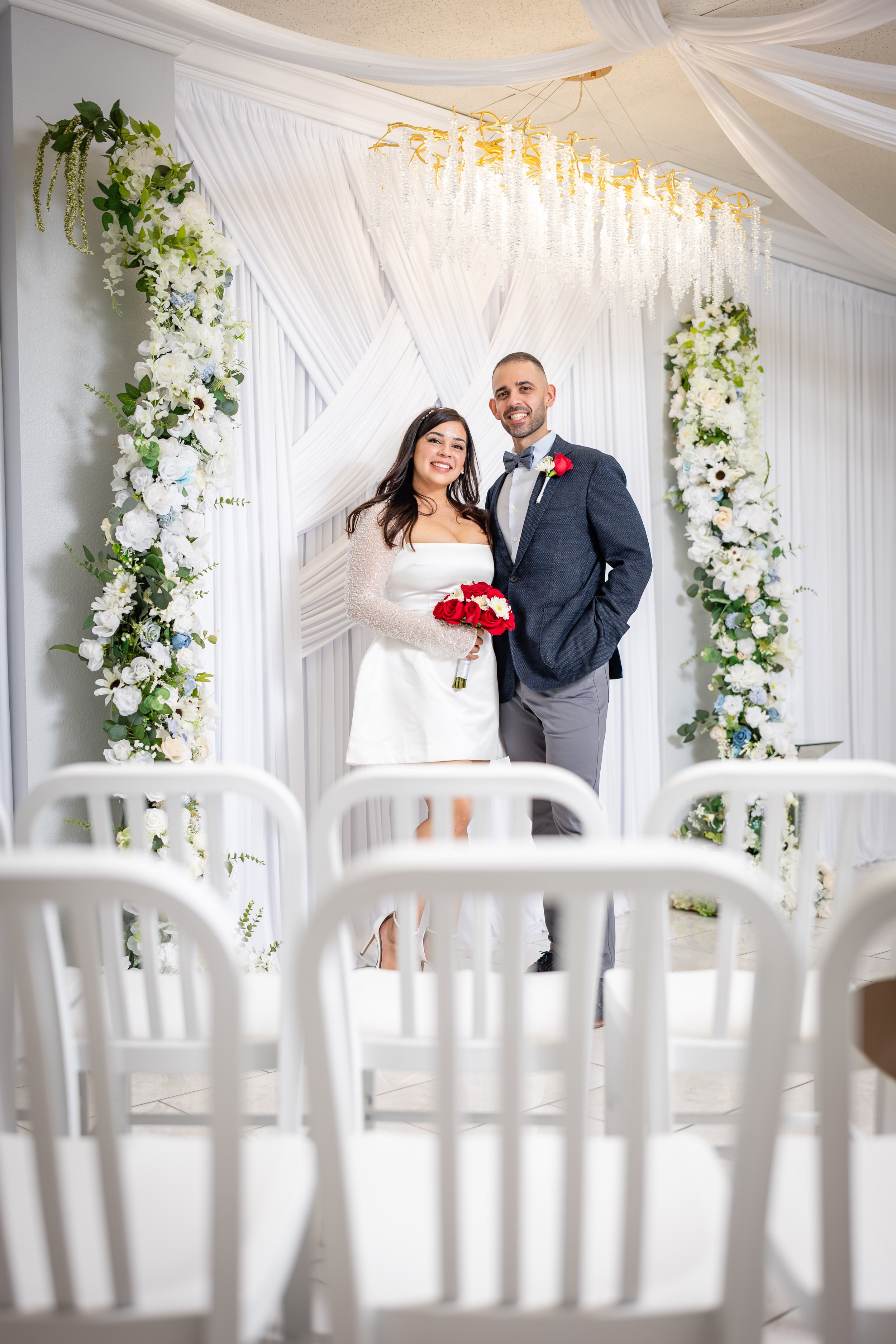 A newlywed couple stands smiling at their wedding ceremony, surrounded by white floral arrangements and draped fabric, with empty white chairs in the foreground.