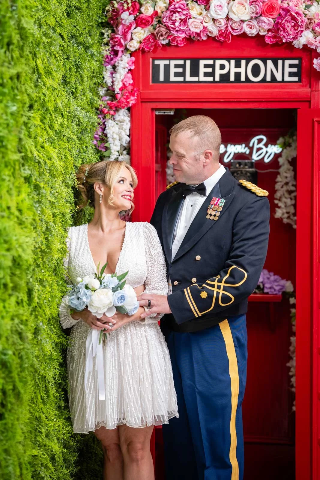A bride and groom standing close together, with the bride holding a bouquet of white and light blue flowers. The groom is dressed in a military uniform with medals, and the bride is wearing a white, knee-length wedding dress. They are in a decorative
