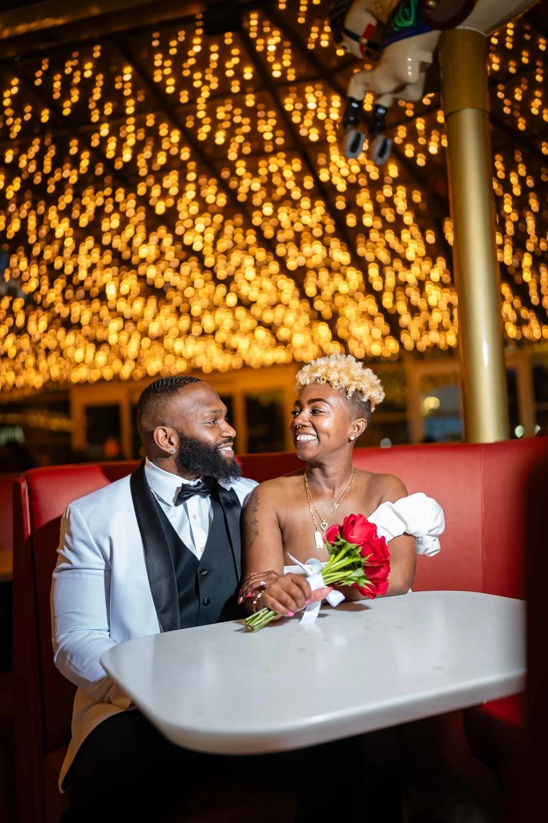 A happy couple, dressed formally, sitting at a table in a decorated venue with warm lighting, the woman holding a bouquet of red roses.