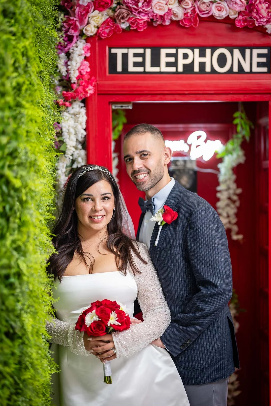 A bride and groom standing together in front of a red telephone booth inside the Say I Do Wedding Chapel in Las Vegas, decorated with flowers, smiling at the camera.