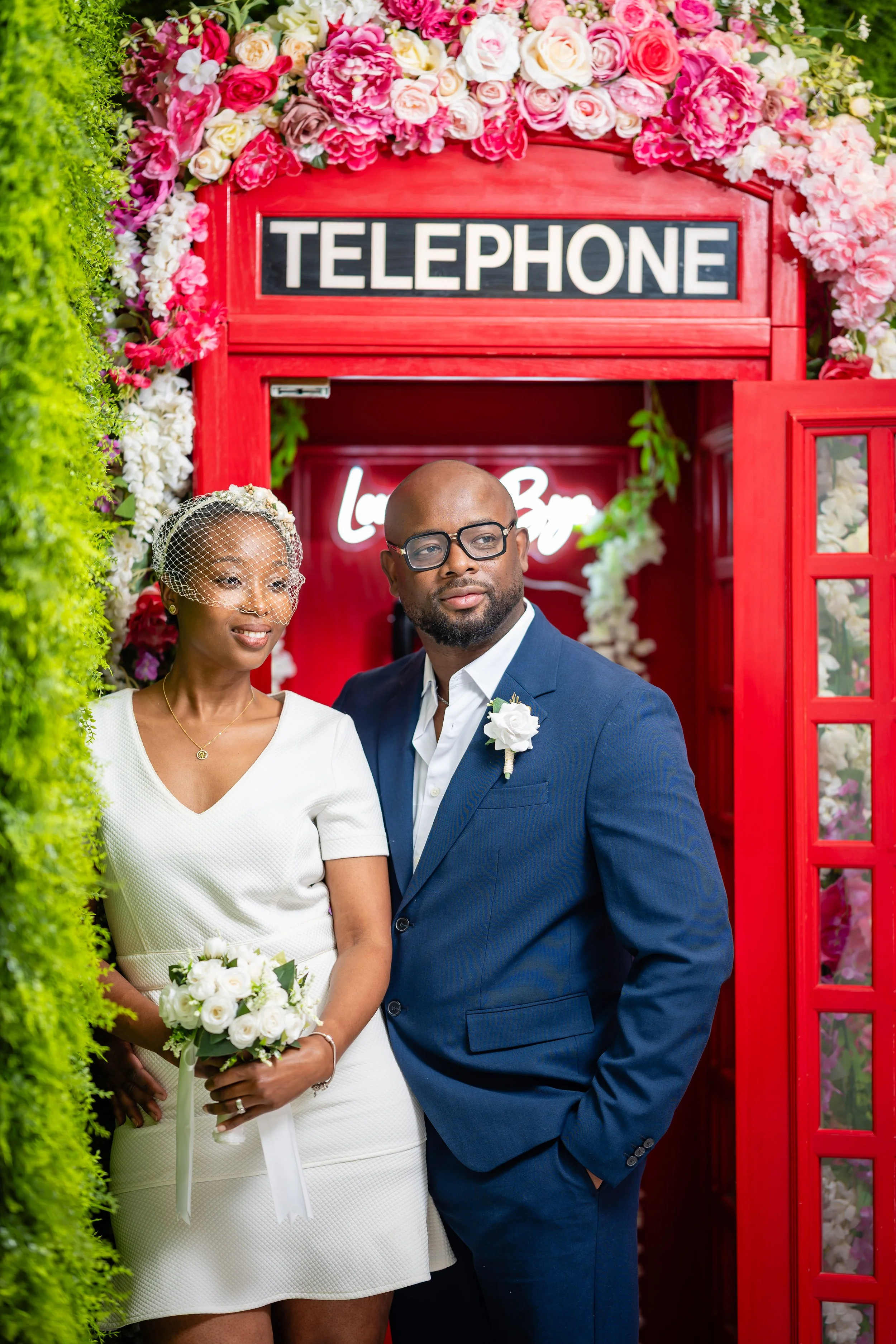 A wedding couple standing in front of a red telephone booth decorated with pink and white flowers. The bride is wearing a white dress with a bouquet of white roses, and the groom is in a blue suit with a white boutonniere. There is a green hedge on t