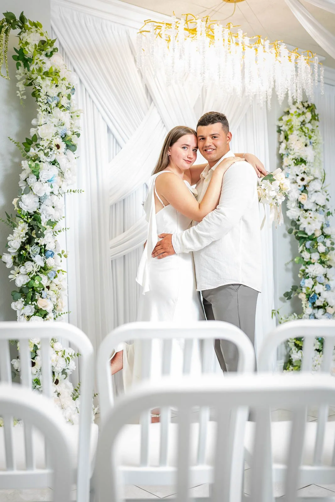 A bride and groom embrace during their wedding ceremony in front of a floral arch with white and blue flowers.