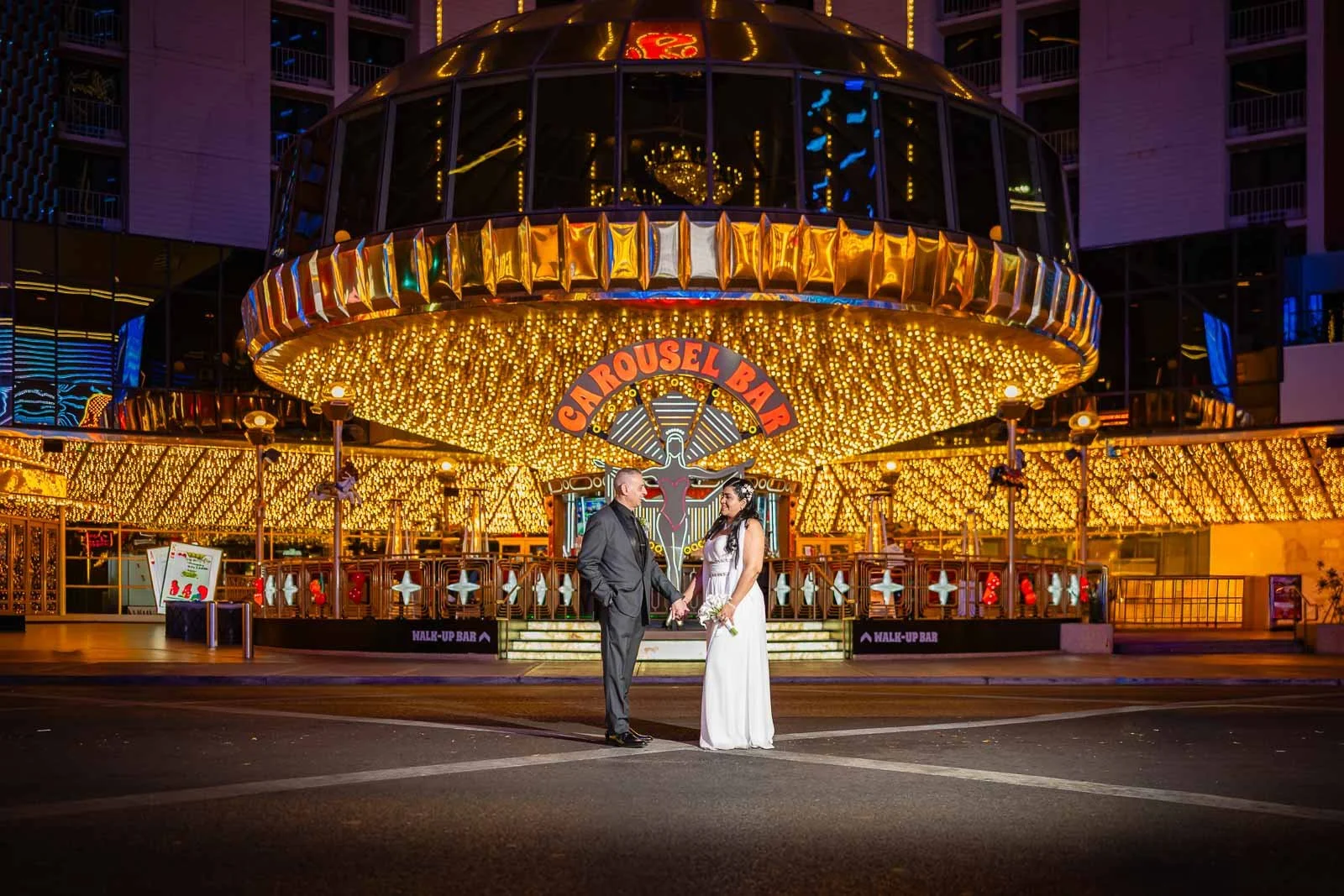 A couple dressed formally, holding hands, standing in front of a brightly lit amusement park ride called Carrousel Bar at night in downtown Las Vegas.