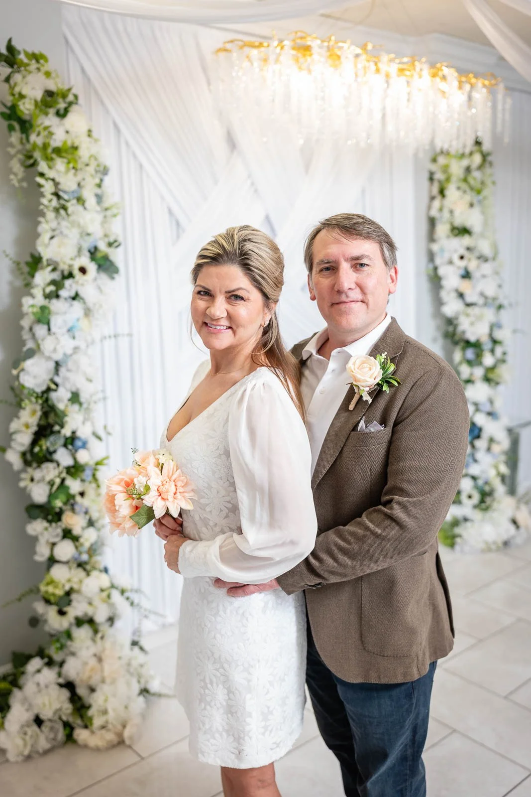 A smiling middle-aged couple dressed in wedding attire posing together at a wedding ceremony, with a floral backdrop and white drapes, at the Say I Do Wedding Chapel in Las Vegas.