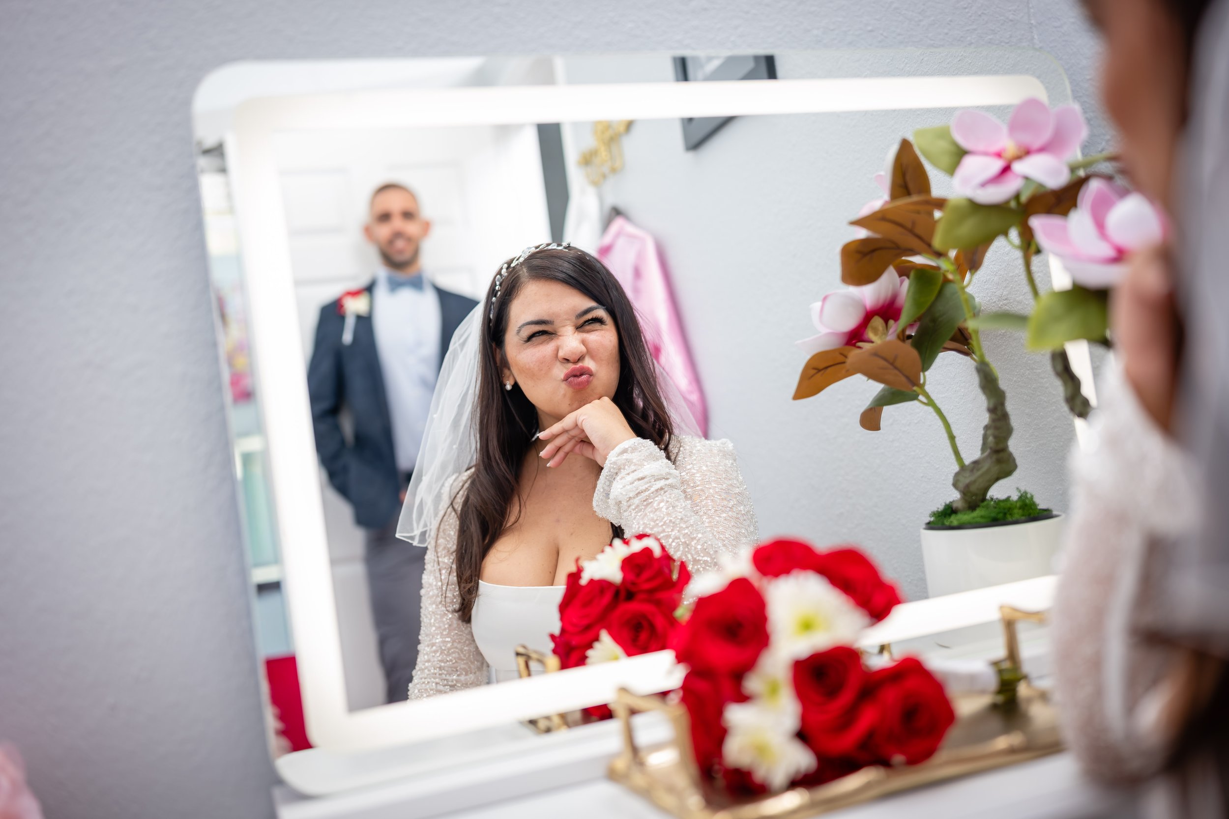 Bride making a silly face at herself in a makeup mirror with wedding bouquet and flowers in the foreground, groom smiling in the background.