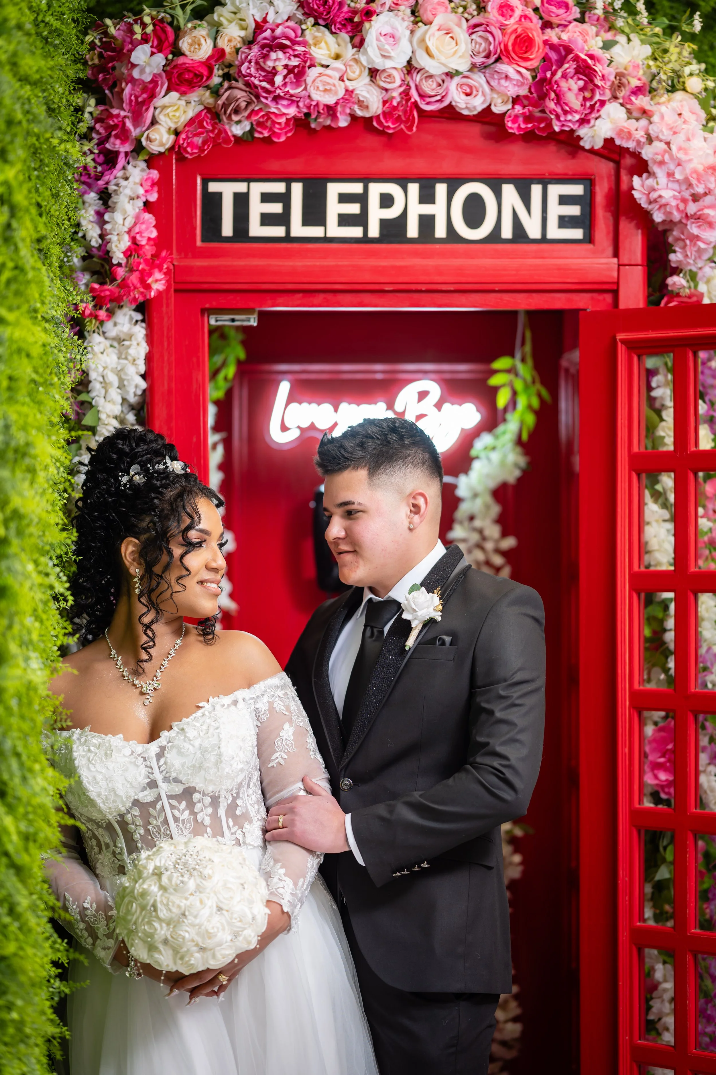 A bride and groom in wedding attire sharing a romantic moment beneath a red telephone booth decorated with pink and white flowers, with a neon sign in the background reading "Love You".