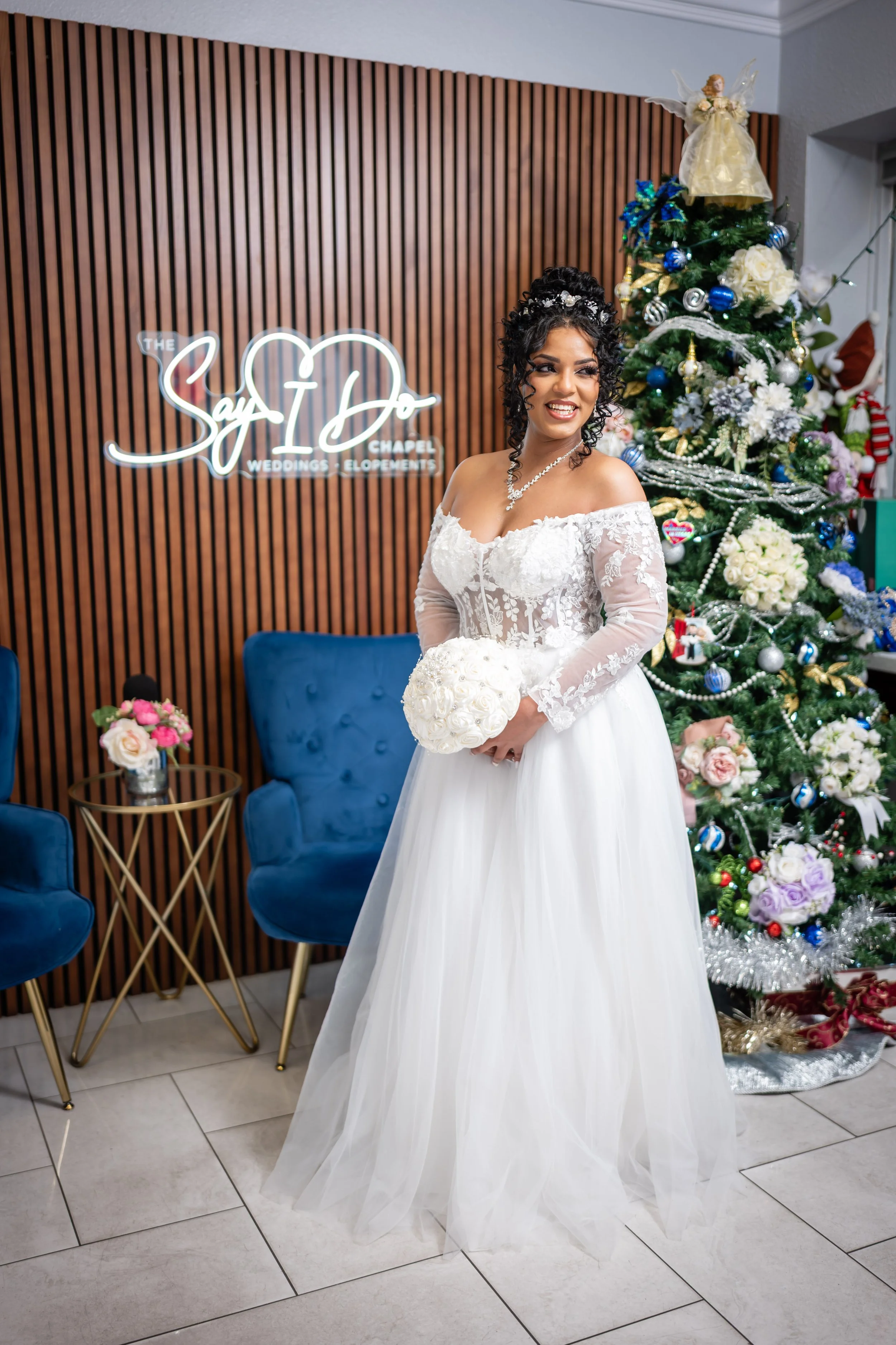 A bride in a white wedding dress holding a bouquet of white roses, smiling indoors near a decorated Christmas tree and a neon sign that reads 'Say I Do Chapel Weddings Elopements'.