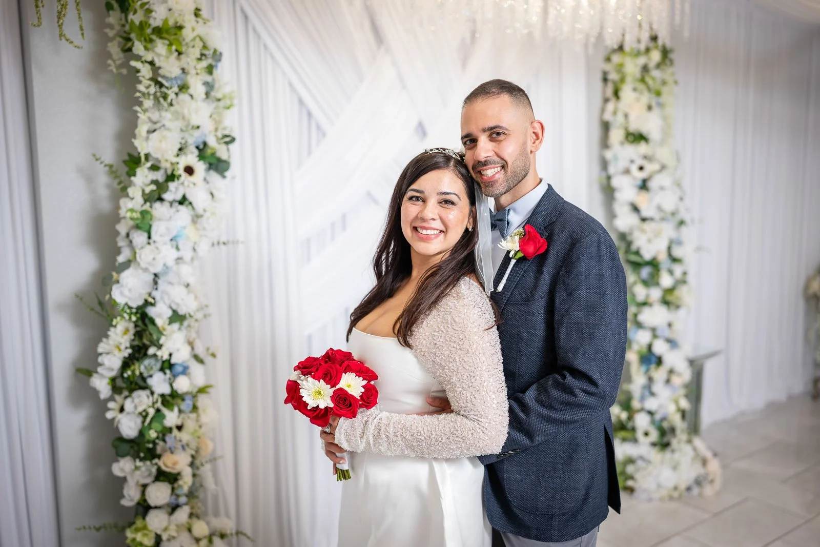 A newlywed couple in wedding attire, smiling and embracing, standing in front of a decorated floral backdrop with white flowers at the Say I Do Wedding Chapel.