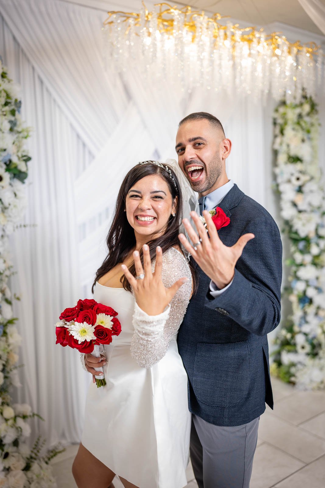 A happy bride and groom showing off their wedding rings at their wedding reception. The bride has a bouquet of red and white flowers. The backdrop features white flowers and drapery with a chandelier above.