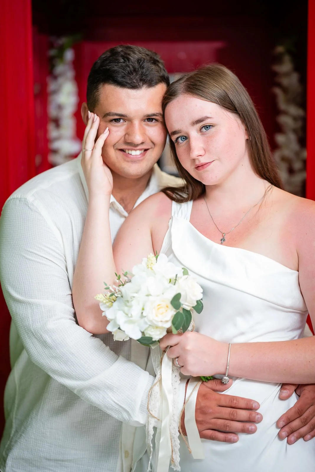 A young couple on their wedding day, with the groom in a white shirt and the bride in a white dress holding a white bouquet of flowers, standing close together in front of a red background.
