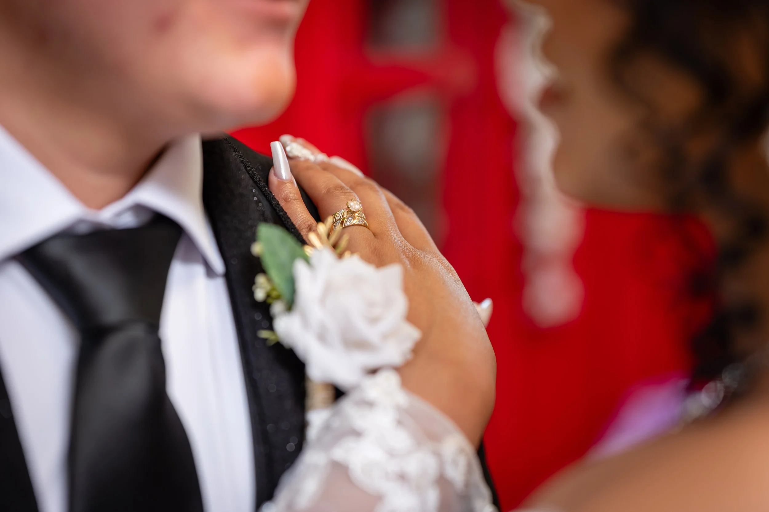 Close-up of a bride and groom during their wedding Ceremony, with the bride's hand on the groom's shoulder showing her wedding rings, with a red background and blurred face details.