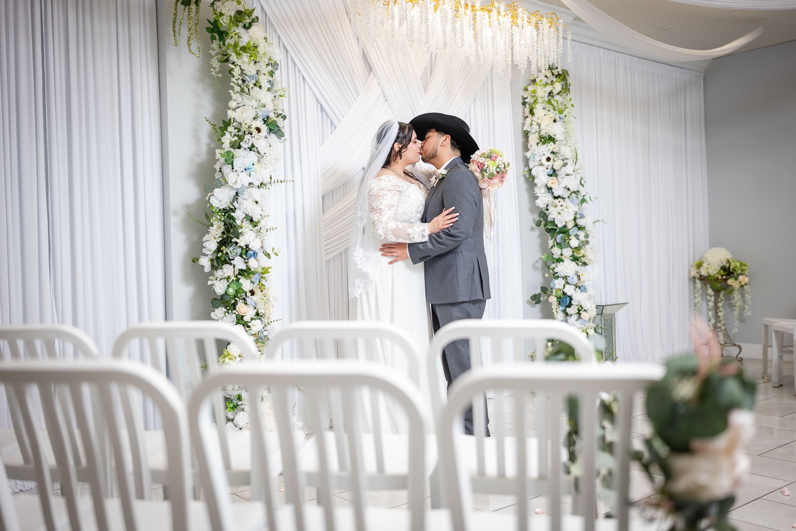 A bride and groom sharing a kiss during their wedding ceremony at the Say I Do Chapel standing under a floral arch decorated with white flowers and greenery, with white draped fabric backdrop.