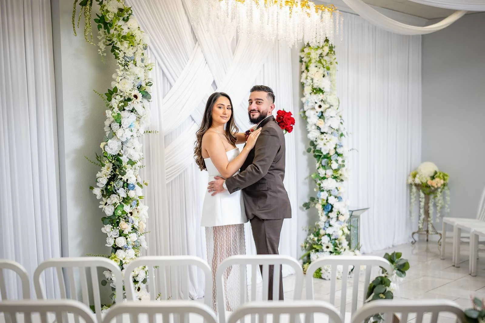 A bride and groom standing close together during their wedding ceremony inside a decorated white venue, with floral arrangements and chairs in the background.