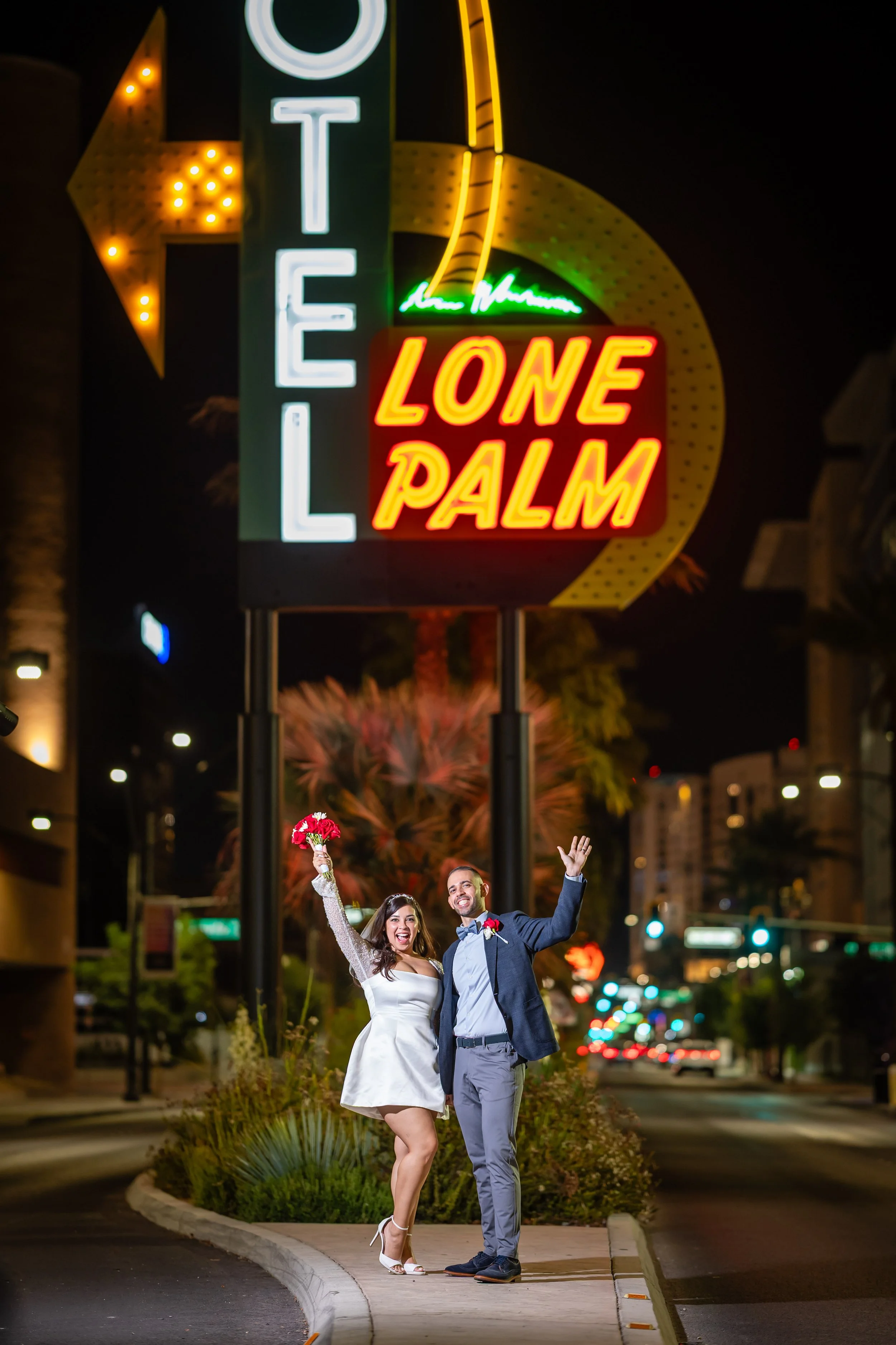 Couple celebrating wedding night in front of neon sign at night, holding bouquet and waving.