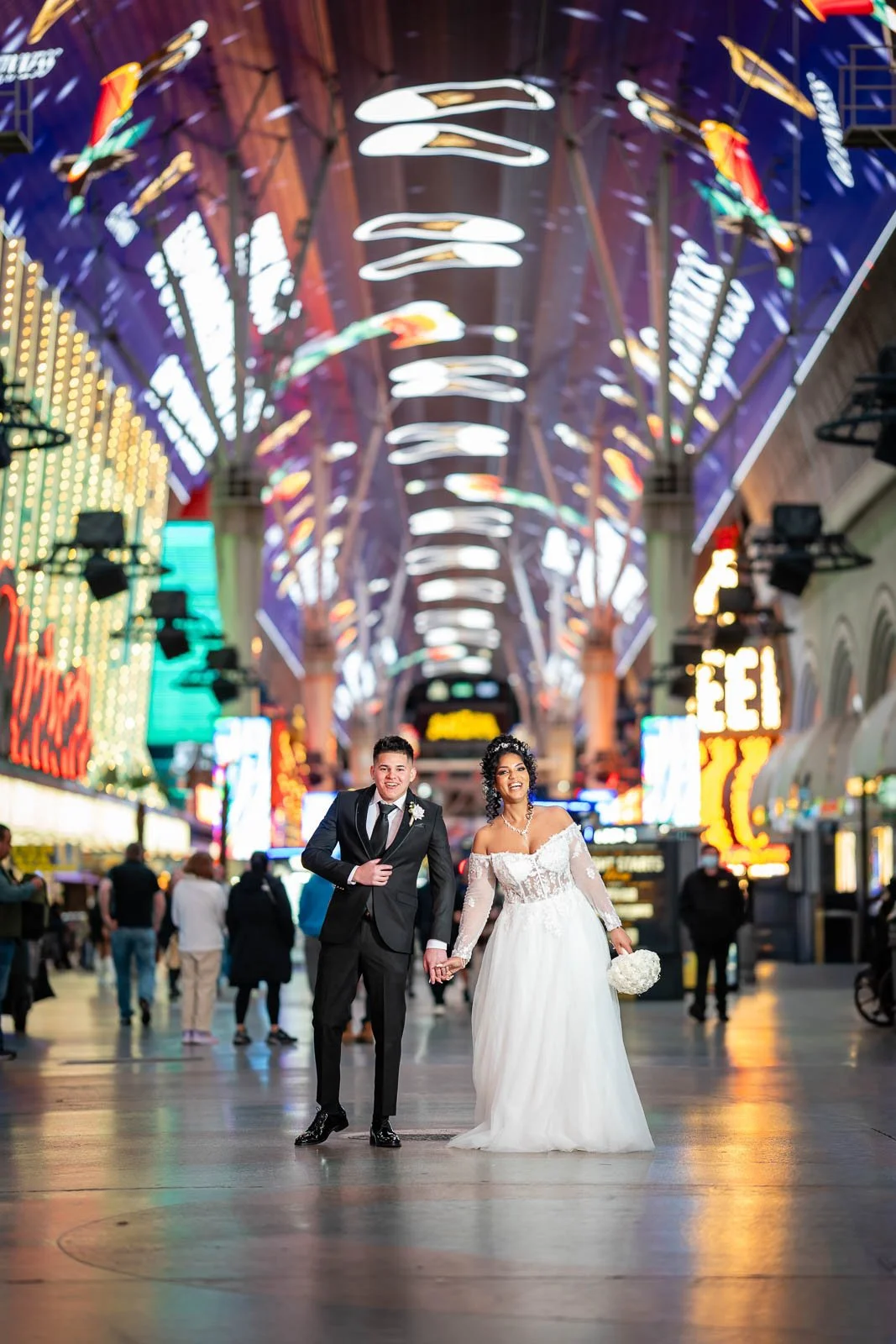 A newlywed couple holding hands and smiling in a brightly lit indoor arcade or entertainment venue, with neon signs and colorful lights in the background.