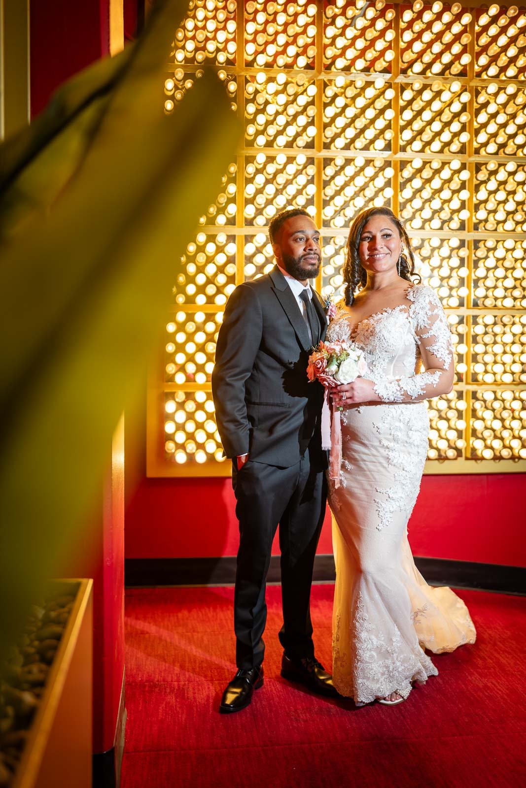 A newlywed couple stands together in front of a decorative wall with warm light bulbs. The bride is in a lace wedding gown holding a bouquet, and the groom is in a black suit with a white shirt and black tie.