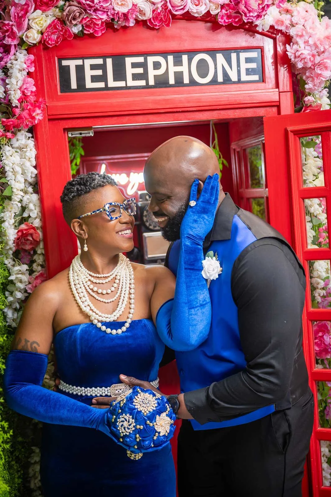 A couple dressed in matching blue and black outfits sharing a romantic moment in front of a red phone booth decorated with pink and white flowers.