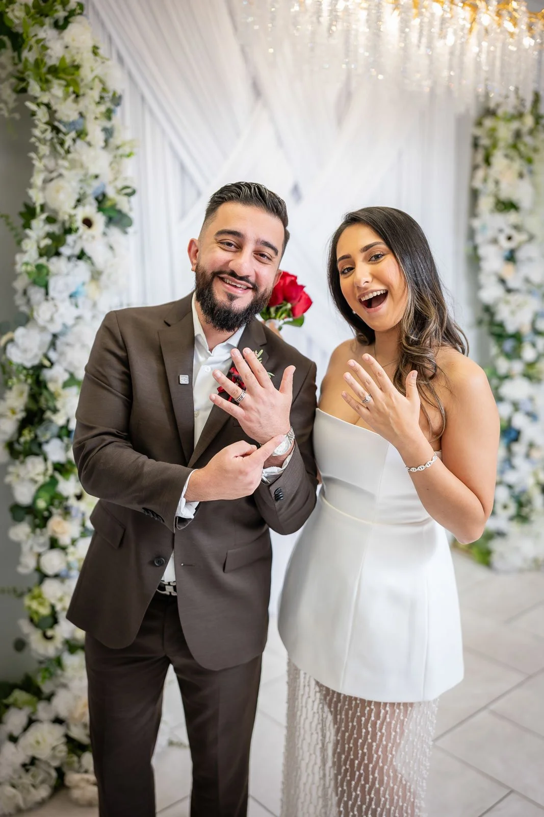A happy couple at their wedding at the Say I Do Chapel, showing off their wedding rings, with floral decorations in the background.