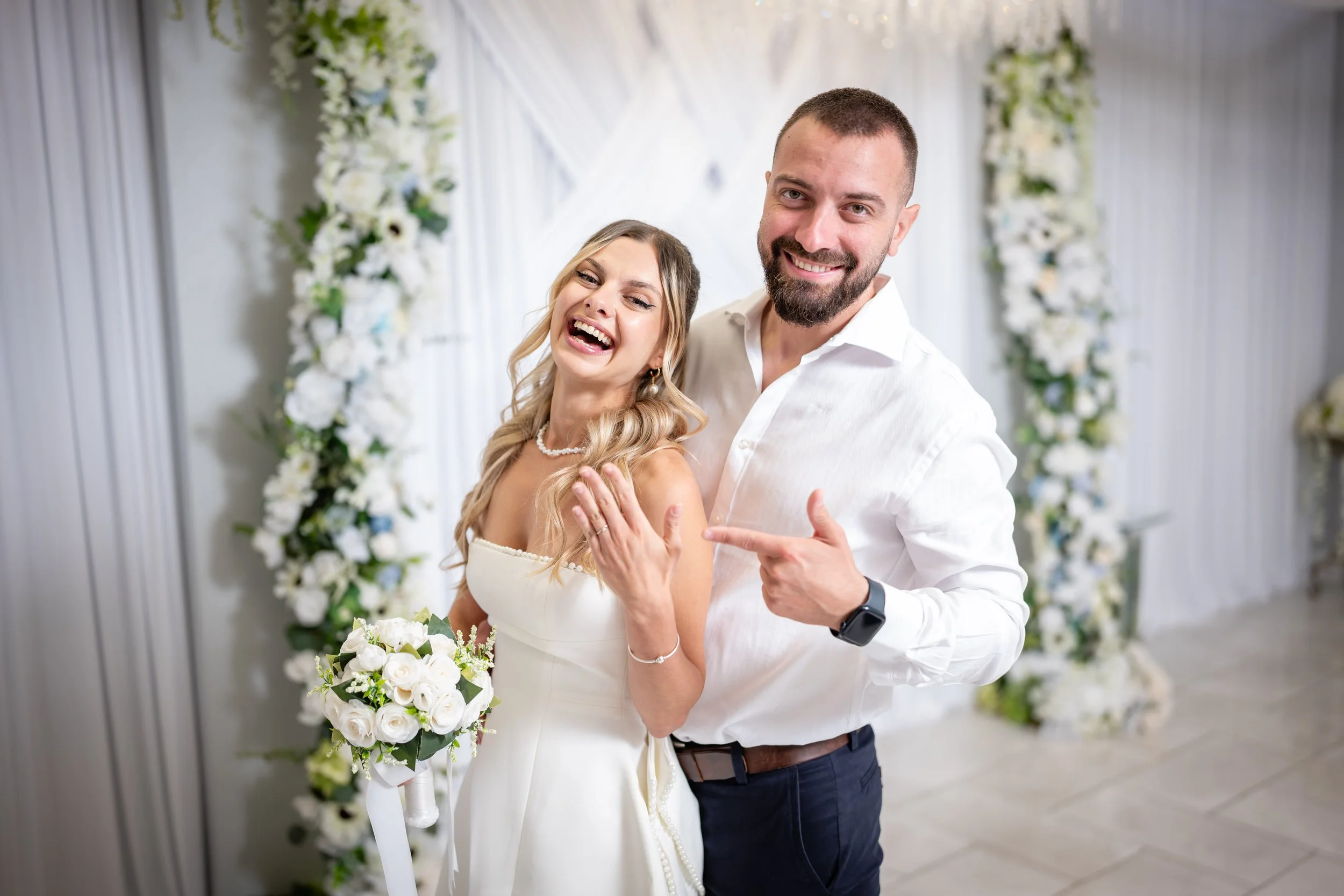 Happy bride holding bouquet of white roses and a smiling groom pointing at her, standing in front of floral wedding arch