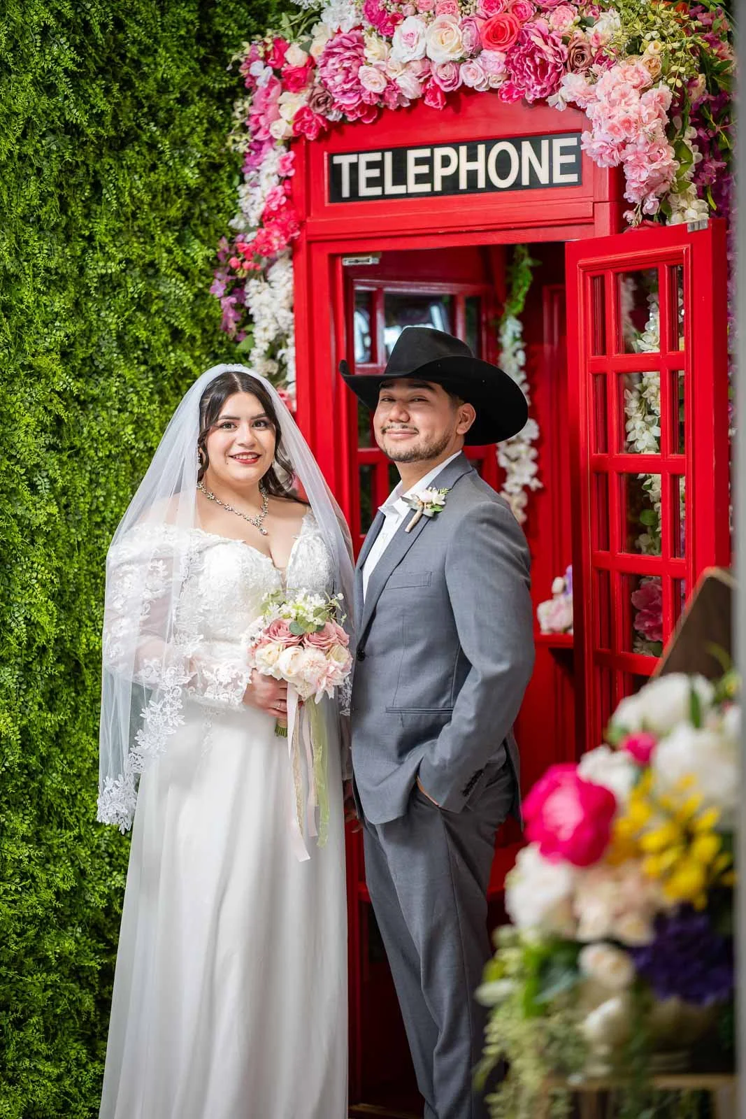 A bride and groom standing in front of a red telephone booth decorated with pink and white flowers. The bride is holding a bouquet of pink and white roses, wearing a white lace wedding dress and veil, smiling. The groom is dressed in a gray suit with