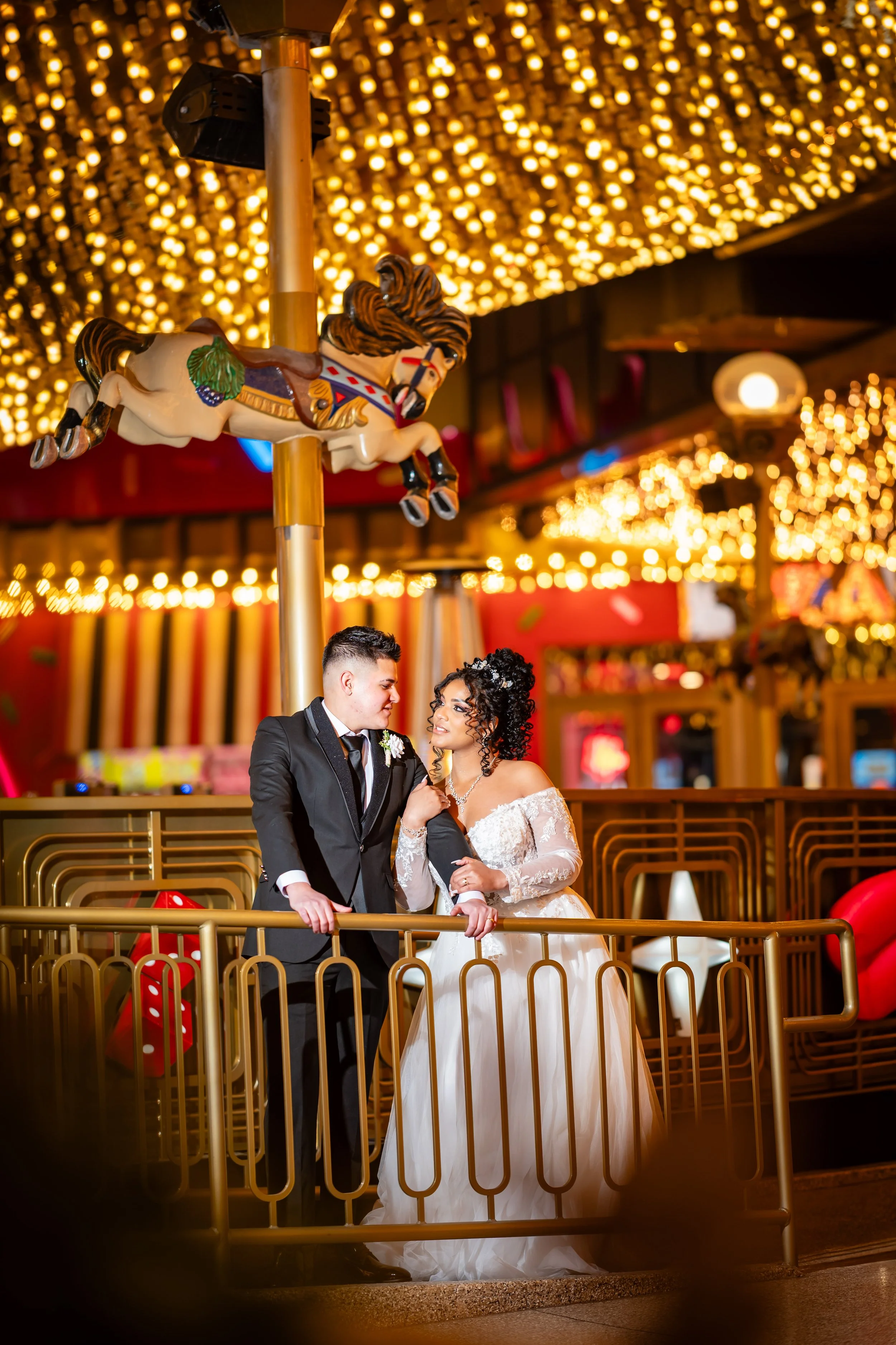 A bride and groom in wedding attire standing inside an amusement park at night with a carousel horse and warm string lights in the background.